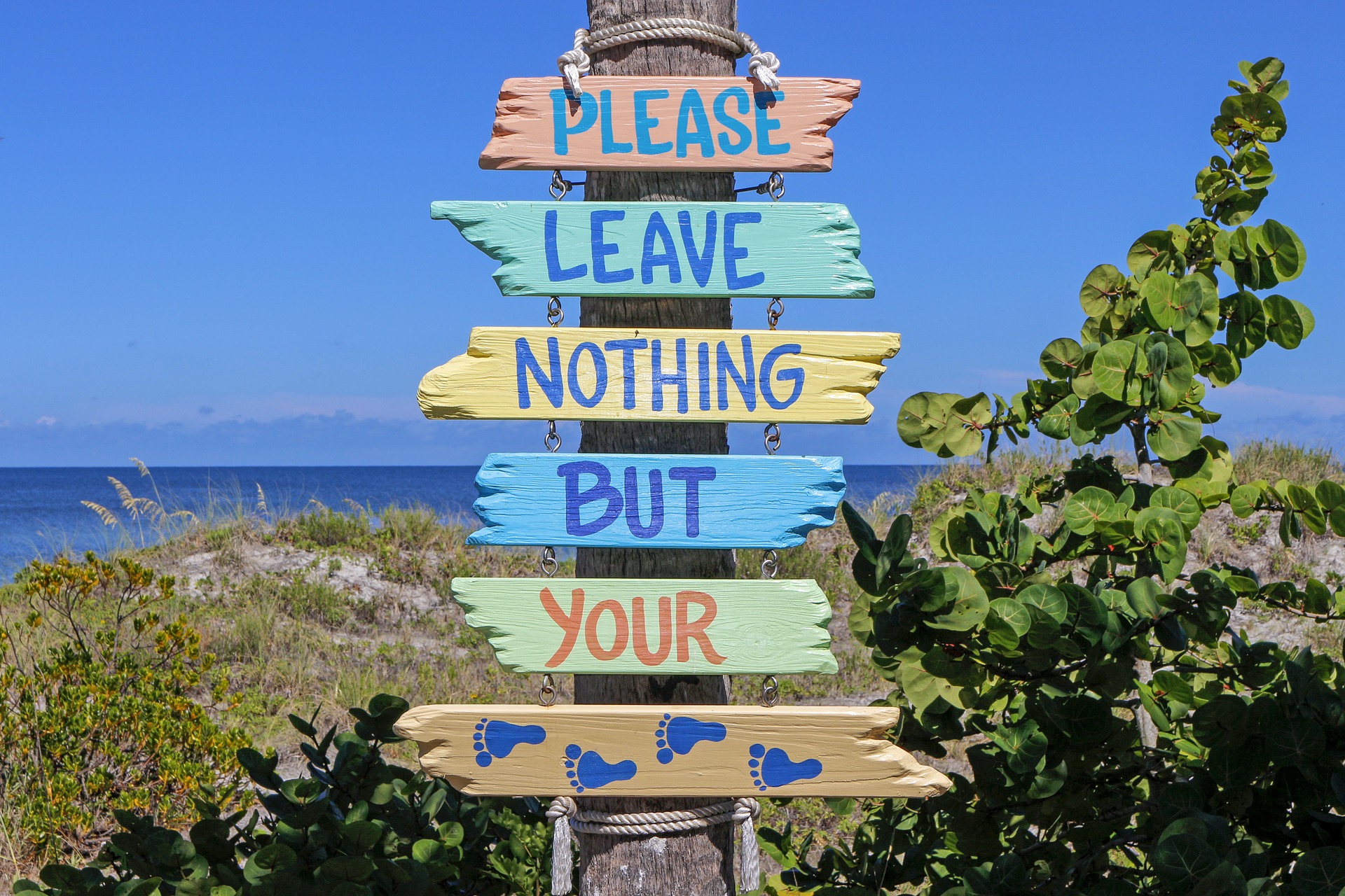 Schild mit der Aufrschirift: "Please leave nothing bur your" der unterste Teil des Schilds zeigt Fußabdrücke. Das Schild ist mitten im Grünen und dahinter sieht man das Meer.