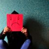 Frau sitzt auf dem Boden und hält sich eine rote Maske mit einem unzufriedenen Smiley vor das Gesicht. Negative Bewertungen – was tun?