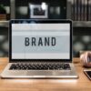 Aufgeklappter Laptop auf dem Brand steht auf Holztisch mit Kaffeebecher, Handy, Kopfhörer
