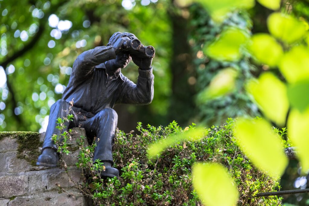 Figur eines Mannes, der auf einer Mauer sitzt, sich herunter beugt und durch ein Fernglas schaut.