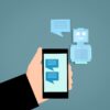 Grafik einer Hand, die ein Smartphone hält. Auf dem Display ein Chat. Im Hintergrund ein Roboter mit Sprechblase.