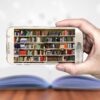 Eine Hand hält ein Smartphone, im Display ist ein volles Bücherregal zu sehen. Im Hintergrund liegt ein aufgeschlagenes Buch auf einem Tisch, aus dem Buchstaben herausfliegen.