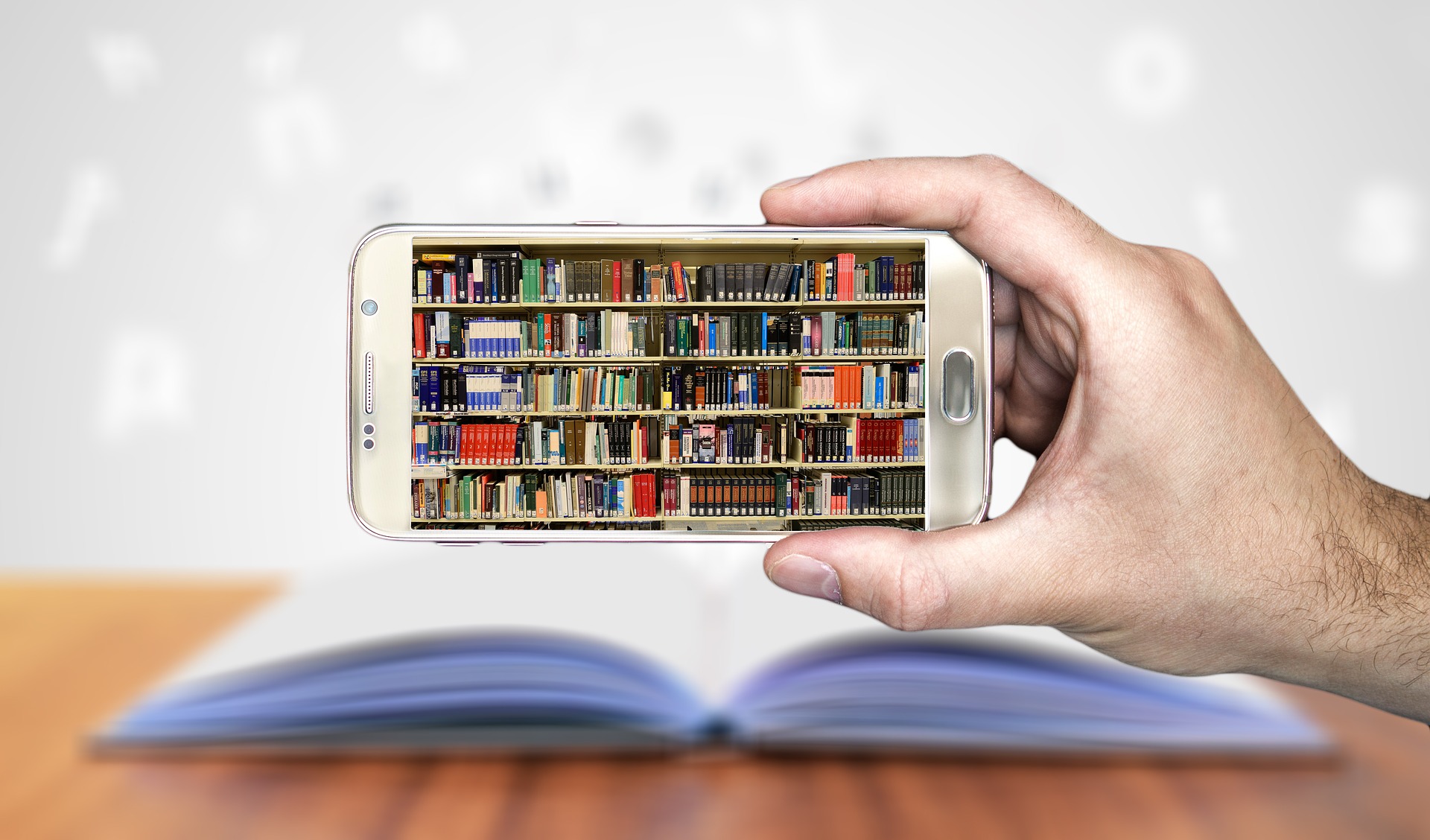 Eine Hand hält ein Smartphone, im Display ist ein volles Bücherregal zu sehen. Im Hintergrund liegt ein aufgeschlagenes Buch auf einem Tisch, aus dem Buchstaben herausfliegen.