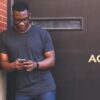 Ein Mann lehnt mit der rechten Schiulter an einer Hauswand und schaut auf sein Smartphone, das er in beiden Händen hält.
