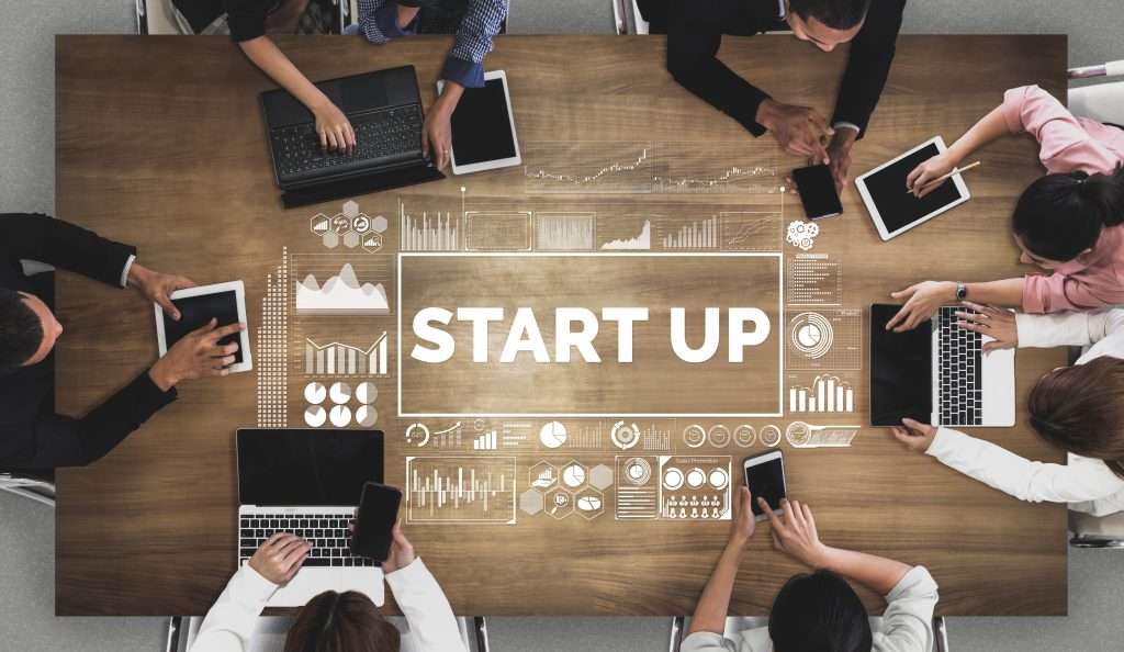 Konferenztisch mit Schriftzug "Start up" in der Mitte, Menschen mit Laptops und Handys sitzen am Tisch