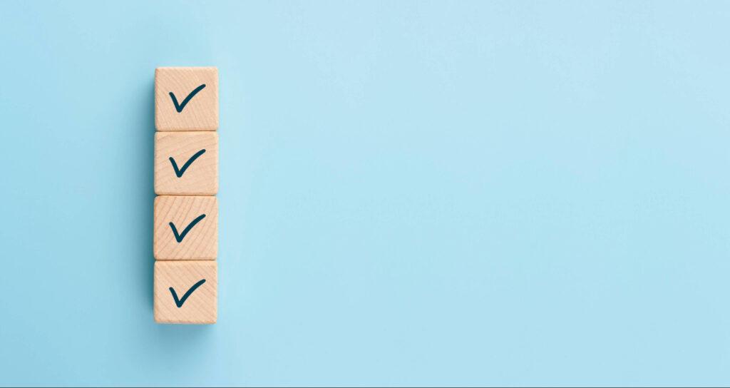 Vier Holzblöcke mit Häkchen als Symbol für erledigte Aufgaben oder Checkliste auf blauem Hintergrund.