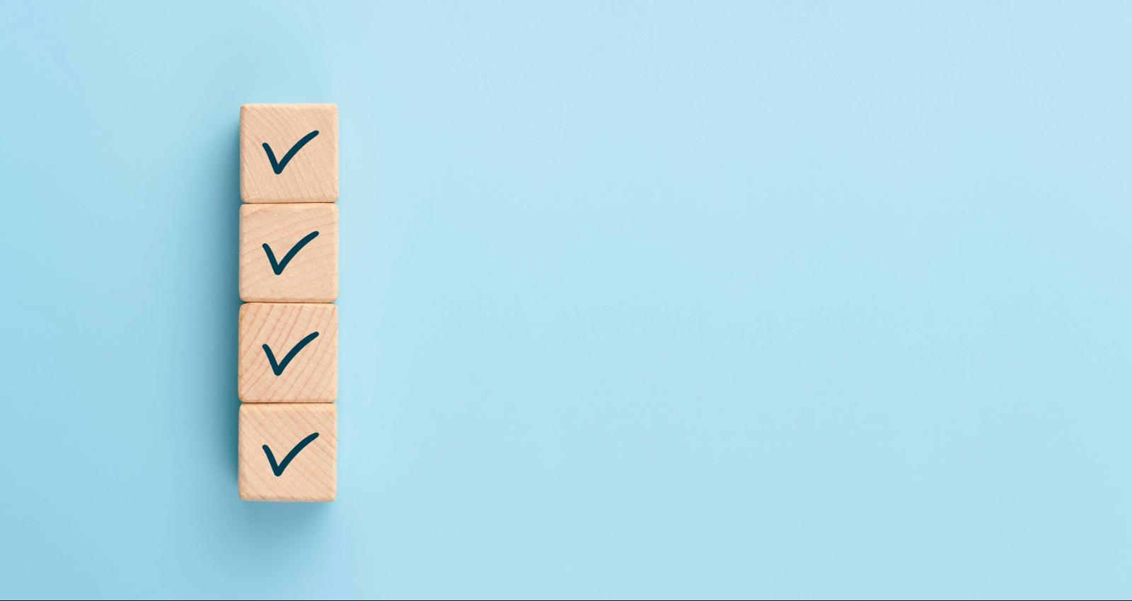 Vier Holzblöcke mit Häkchen als Symbol für erledigte Aufgaben oder Checkliste auf blauem Hintergrund.
