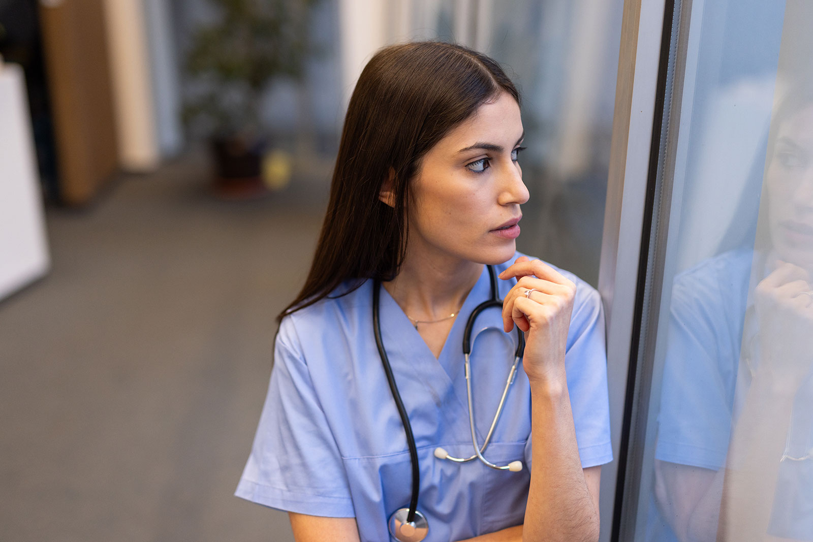 Nachdenkliche medizinische Angestellte in blauer OP-Kleidung und mit Stethoskop, die nachdenklich aus dem Fenster im Krankenhausflur schaut. 